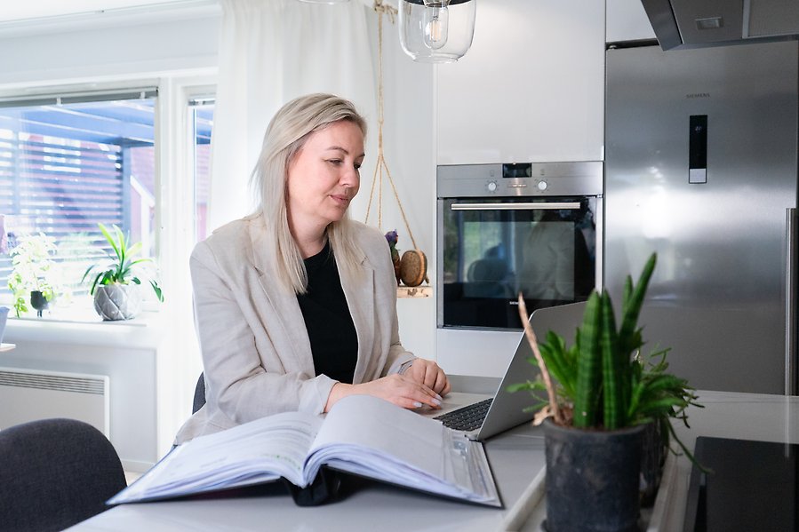 Kvinna sitter i köket med sin dator och med en pärm uppslagen.