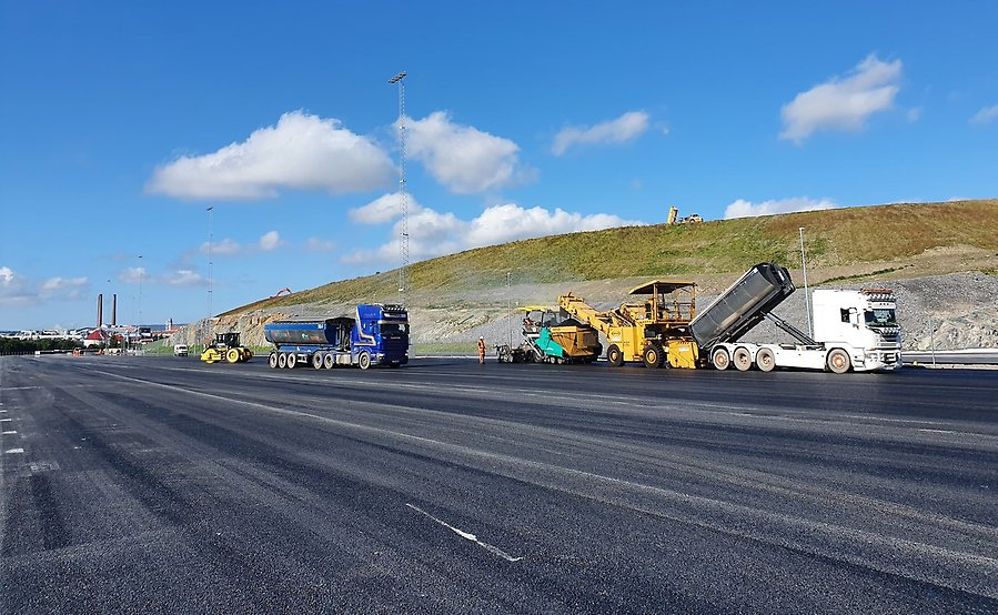 En blå och en vit lastbil samt en gul vägvält lägger ut asfalt på ett stort öppet område i logistikparken