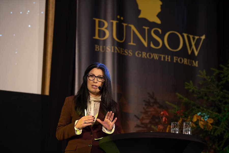 Moderator Anna Olin Kardell på en stor svart scen. På väggen bakom står det med stor guldig text "Bünsow Business Growth Forum". 