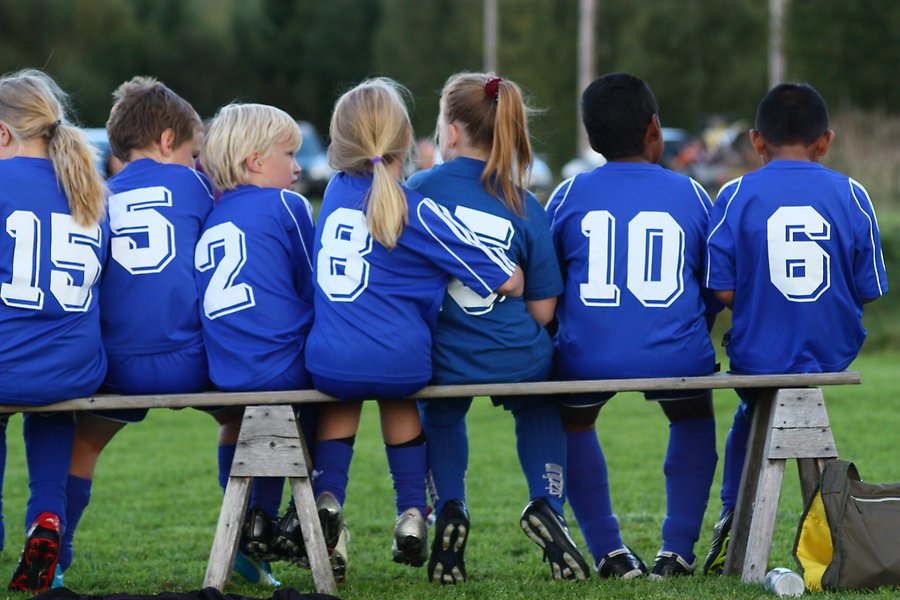 Flera barn sitter på rad på en träbänk. De har på sig blå fotbollströjor och blåa shorts. På ryggarna har alla ett nummer i vit färg.