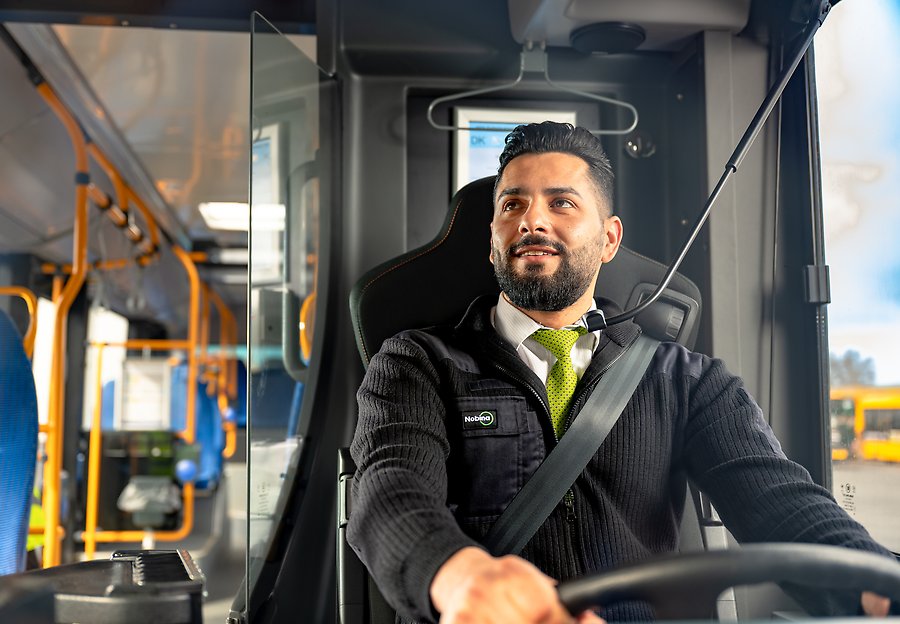 En manlig bussförare sitter bakom ratten i en lång stadsbuss. Han kollar i backspegeln och är klädd i svarta kläder och har en grön slips runt halsen.