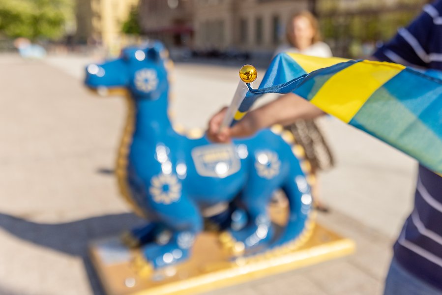 I framgunden syns en hand som håller i en svensk flagga. I Bakgrunden syns en staty som föreställer en drake. Draken är blå med gulddetaljer. 