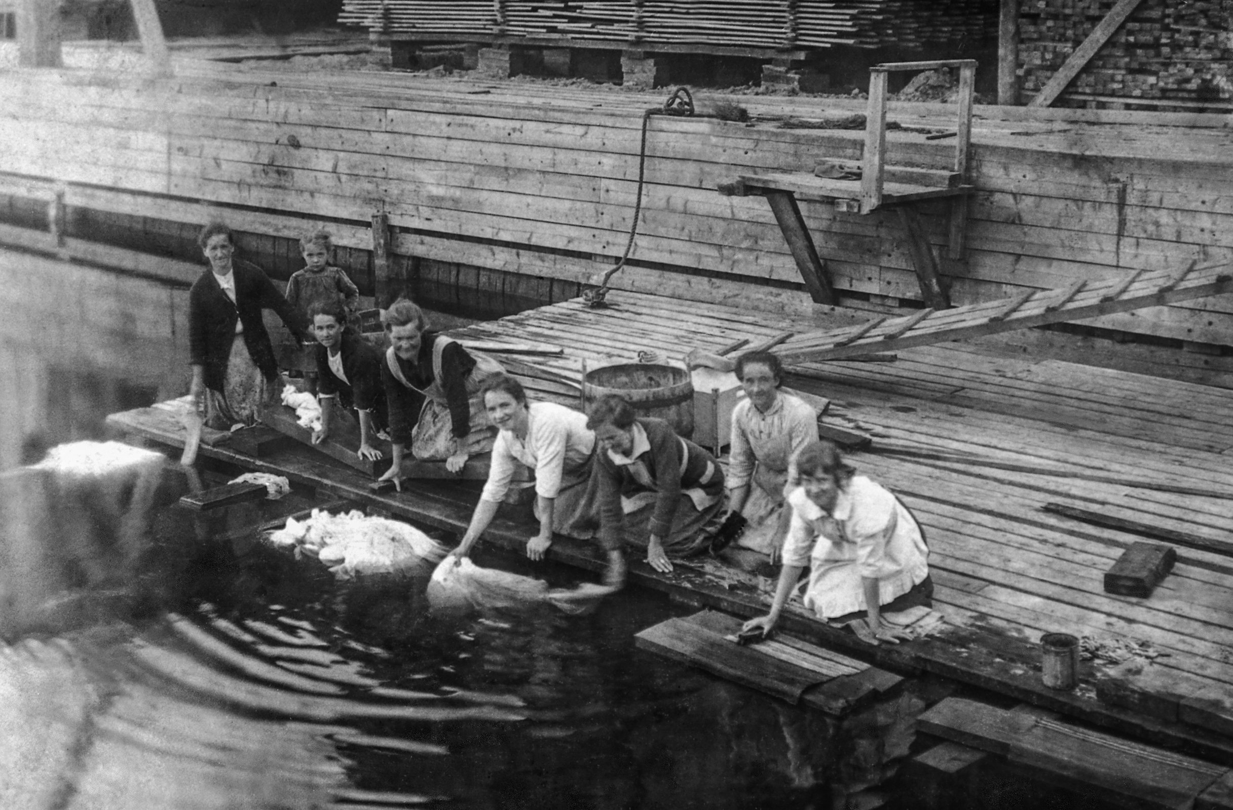 Ett barn och sju kvinnor tvättar vid sågverkskajen, runt 1920-1940.