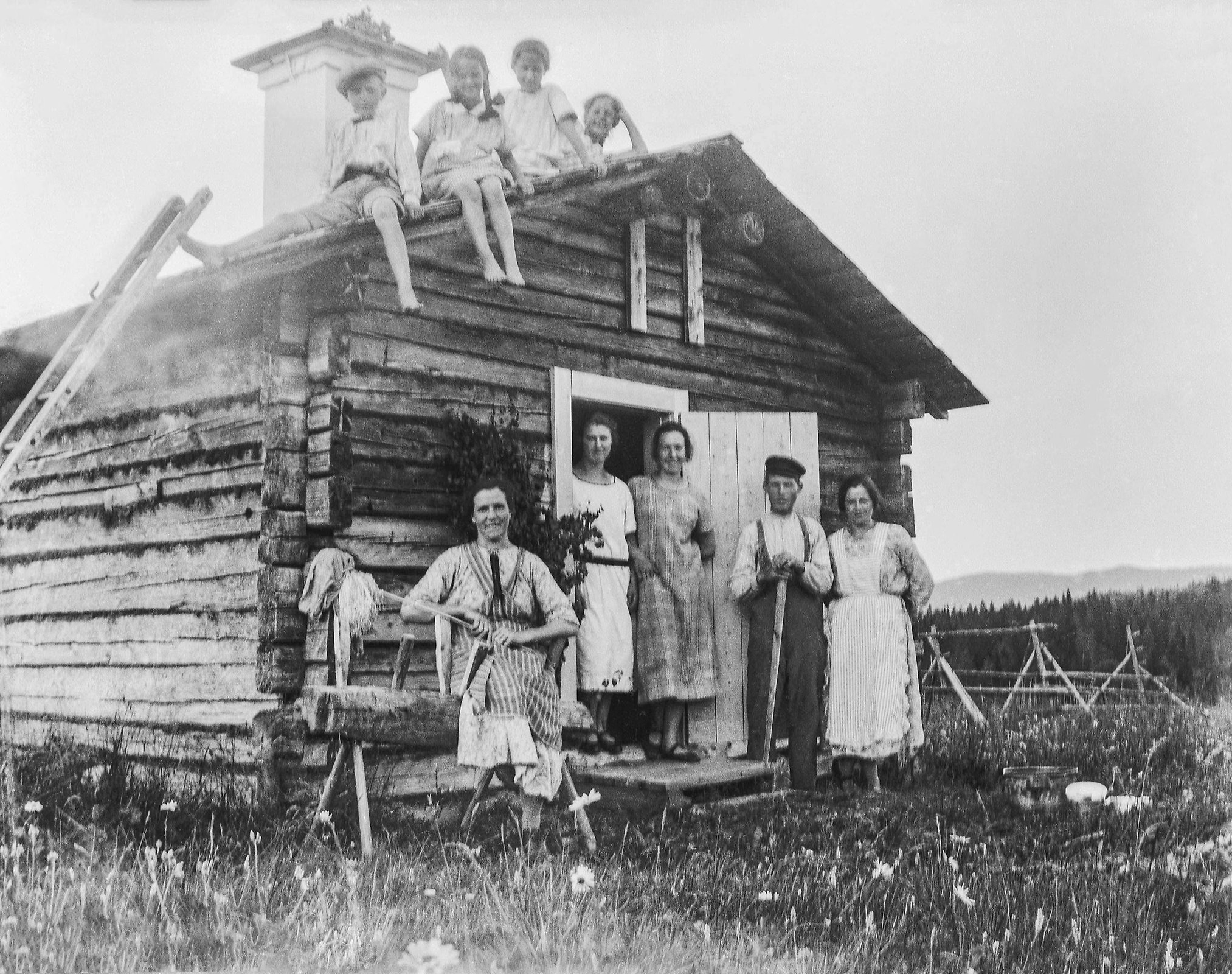Höglunds stuga vid Fanbybodarna. Framför stugan sitter en person och fyra står. På taket sitter fyra barn, runt 1923-25.