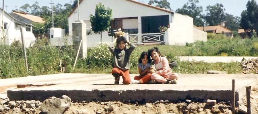 Foto. Tre barn sitter på en betongplatta, grunden till ett hus.