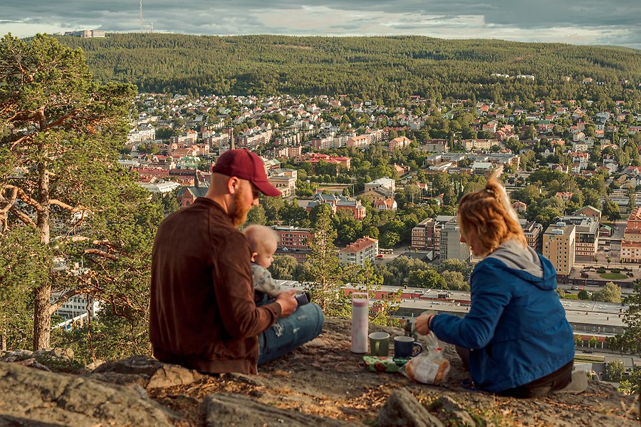 En kvinna och en man sitter med ett litet barn på en klippa och fikar. Man ser mycket skog och en stad i sten.