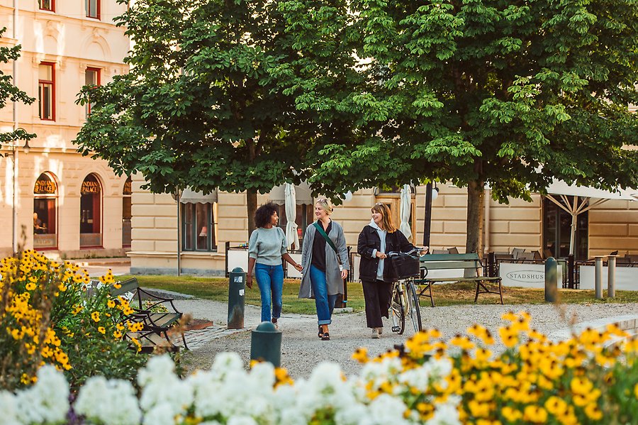 Tre kvinnor går i en park i Sundsvall. De alla är glada och är på väg mot ett okänt mål. Runt omkring dem står det mycket blommor i gult och vitt och grönt.
