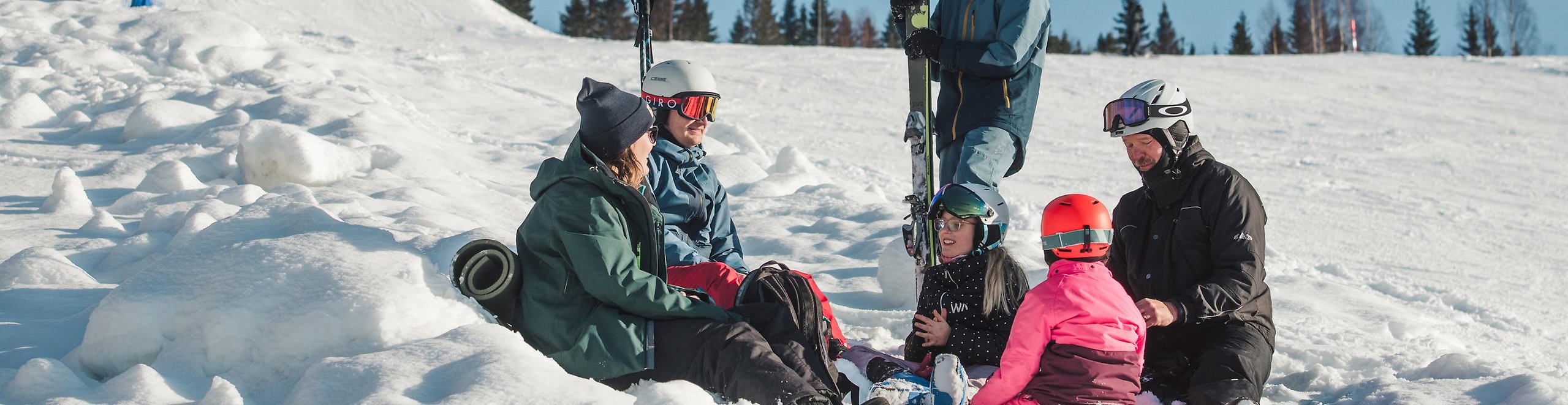 Vuxna och barn med slalomutrustning sitter på liggunderlag i snön.