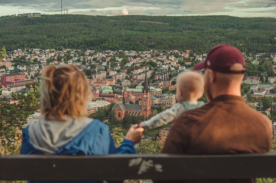En kvinna och en man sitter på en bänk tillsammans med ett barn på Norra Berget. De blickar ut över Sundsvalls stenstad, all natur och Södra Berget.